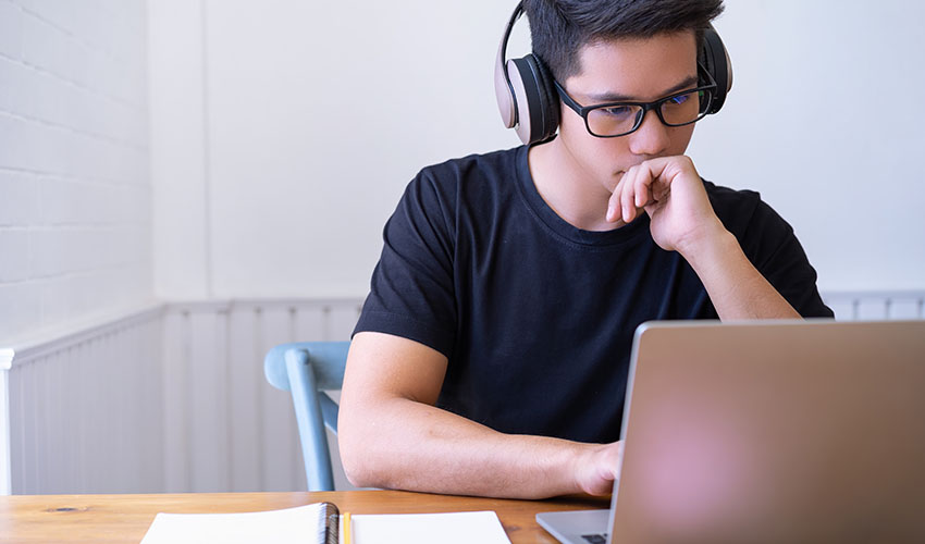 A man with glasses and headphones is focused on his laptop, engaged in work or study