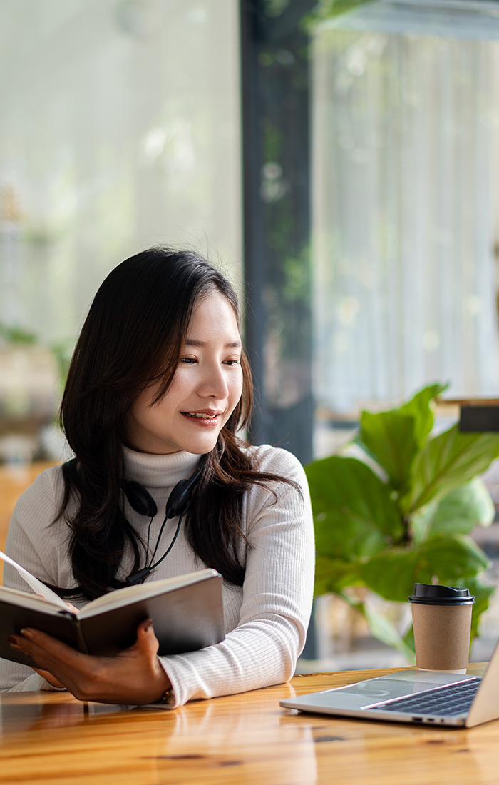 A CSUN student, an Asian woman, joyfully reads a book at a table, with her laptop positioned next to her.