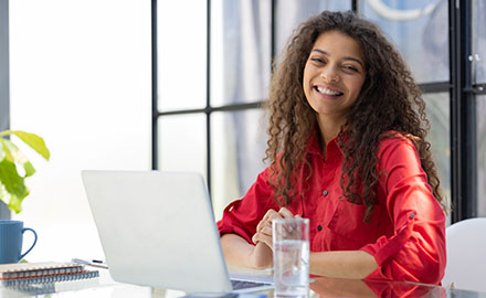 A smiling woman sits at a table with her laptop, engaged in an online course as a dedicated student.