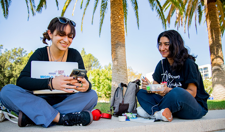 Two CSUN students seated on the ground, engaged with their laptops, collaborating on academic work outdoors.